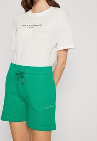 Bílý bavlněný tričko s logem "TOMMY HILFIGER", kombinované s zelenými bavlněnými šortkami s šňůrkou a detailem loga blízko lemu.