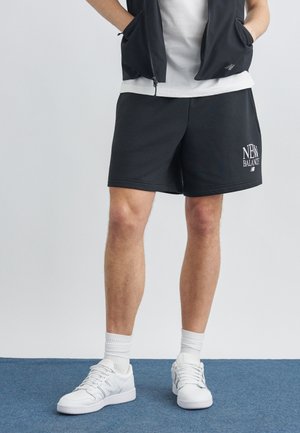 Person iført sorte New Balance-shorts, hvide sneakers, sokker, en hvid t-shirt og en sort lynlåsvest med hænderne i lommerne.