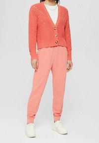 Koraal geknoopte cardigan met V-hals en drie houten knopen, gecombineerd met roze sweatpants met elastische manchetten en zijzakken. Witte sneakers.