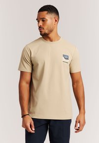 Beige kortärmelig T-shirt gemaakt van zacht stof, met een klein blauw-wit grafisch logo op het linkerborstgebied.
