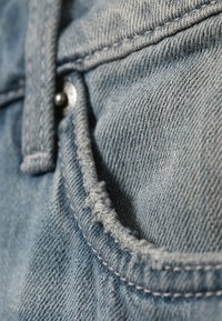Ύφασμα denim σε ανοιχτό μπλε με ελαφρώς φθαρμένη άκρη. Κοντινή λήψη δείχνει λεπτομέρειες ραφής και μεταλλικό κουμπί στην τσέπη.