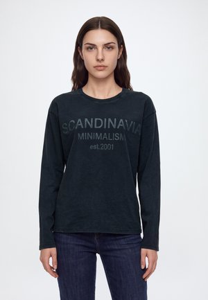 Une femme porte un haut foncé à manches longues avec l’inscription « Scandinavian Minimalism est. 2001 » et un jean foncé, sur un fond blanc uni.