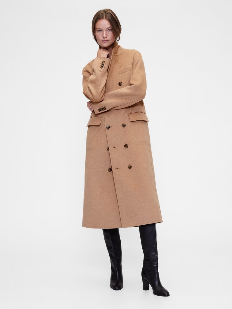 Manteau en laine beige croisé avec de gros boutons, col à revers, poche avant, et longueur jusqu'au genou, associé à des bottes noires montant jusqu'aux genoux.