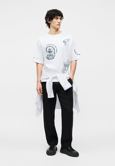 Νέος άνδρας με κατσαρά μαλλιά, που φοράει λευκό μπλουζάκι με σχέδιο, μαύρο παντελόνι, μαύρα αθλητικά παπούτσια και λευκό πουκάμισο δεμένο στη μέση του.