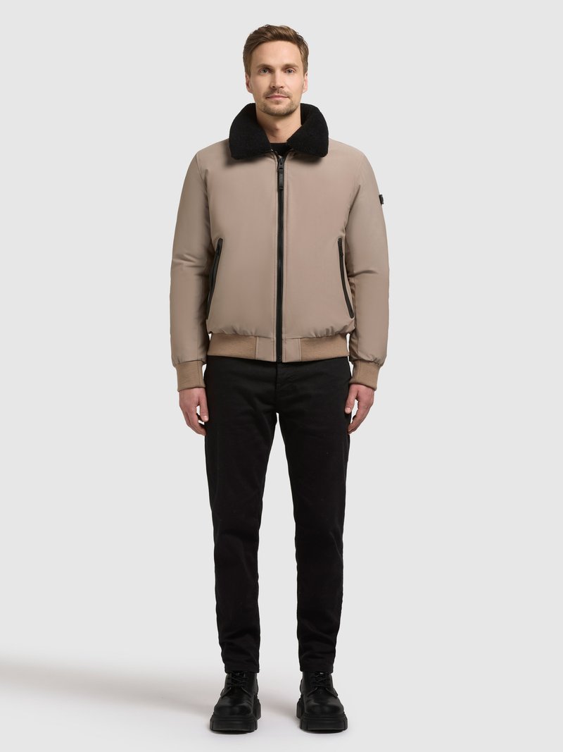 Veste aviateur beige avec col en fausse fourrure noire, poches latérales zippées, poignets et taille côtelés, portée avec un pantalon noir et des chaussures noires.