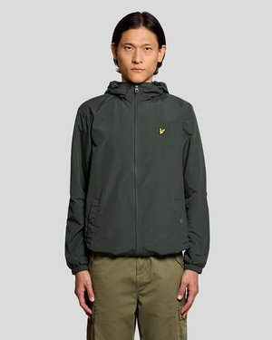 Homme portant une veste à capuche zippée vert foncé avec un logo jaune sur la poitrine et un pantalon cargo olive, debout devant un fond clair uni.