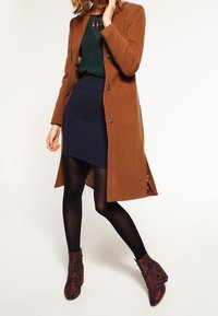 Manteau en laine marron avec boutons, haut vert, jupe côtelée marine, collants noirs et bottines à motifs dans des tons bordeaux et bleu foncé.