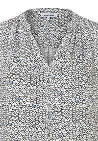 Weiße Bluse mit einem sich wiederholenden Muster aus schwarzen und blauen Herzen, mit V-Ausschnitt und gerafften Details an den Schultern.