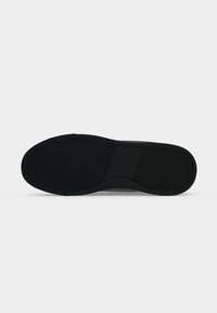 Semelle de chaussure noire avec motif antidérapant texturé et partie médiane lisse sur fond gris clair uni.