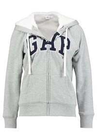 Sudadera con cremallera de color gris claro, con forro de felpa blanca, capucha con cordones blancos, bolsillos frontales y "GAP" en letras grandes color azul marino en el pecho.