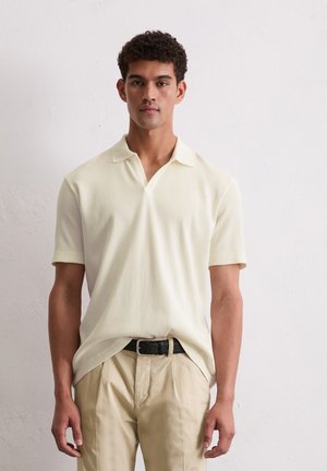 Polo-shirt in lichtcrème met geribbelde structuur, korte mouwen en een V-halskraag. G gedragen met beige broeken en een zwarte riem.