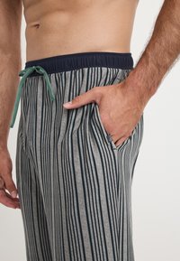 Grijze loungebroek met een marineblauwe elastische tailleband, groene trekkoord en verticale marineblauwe strepen. Bevat een zijzak. Zachte stof.