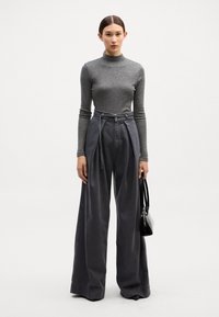 Top de manga larga gris con cuello alto, combinado con pantalones oscuros de pierna ancha y cintura alta de color gris. La modelo sostiene un pequeño bolso negro.