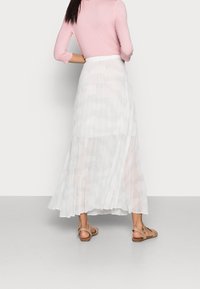 Donna che indossa una lunga gonna plissettata bianca trasparente, un top rosa chiaro a maniche lunghe e sandali beige aperti che sta in piedi con le mani sui fianchi.