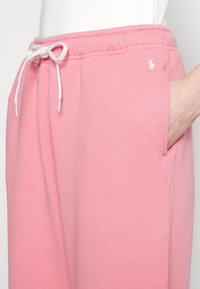 Světle růžové tepláky vyrobené z měkké látky, s elastickým páskem na stažení, bočními kapsami a malým bílým logem.