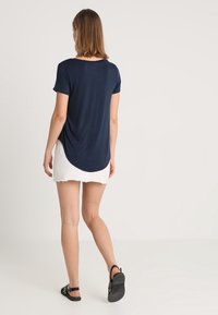 Kvinna som går bort, klädd i en marinblå kortärmad skjorta, vit fransad minikjol och svarta nitar sandaler, mot en enfärgad ljus bakgrund.