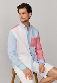 Uomo che indossa una camicia a maniche lunghe a blocchi di colore in azzurro, bianco e rosso, seduto su uno sgabello di legno davanti a uno sfondo grigio uniforme.