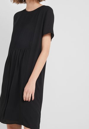 Kobieta w czarnej sukience z krótkim rękawem i plisowanym przodem, stojąca na jednolitym, jasnoszarym tle.