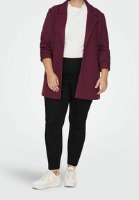 Blazer bordeaux avec un design structuré, manches retroussées, porté sur un haut blanc à côtes et un pantalon noir skinny, associé à des baskets blanches.