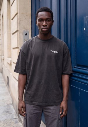 Tânăr cu păr negru și scurt, purtând un tricou larg, închis la culoare, cu textul „Dropsize”, stând lângă o ușă albastră pe trotuarul unui oraș.