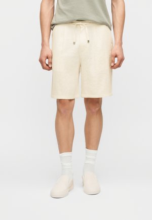 Άνδρας που φοράει μπεζ σορτς με κορδόνι, ανοιχτό πράσινο μπλουζάκι, λευκές κάλτσες και μπεζ παπούτσια τύπου slip-on σε λευκό φόντο.