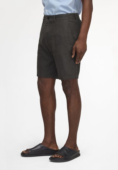 Mann trägt schwarze Shorts, hellblaues Hemd mit Knöpfen und schwarze Slides-Sandalen, steht vor weißem Hintergrund.