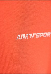 Felrood oranje stof met een gladde textuur. Het "AIM'N SPORT"-logo in het wit, prominent op het oppervlak geplaatst.