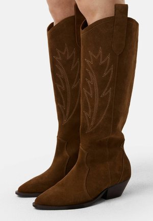 Bottes de cow-boy en daim marron, arrivant au genou, avec des orteils pointus et un talon empilé. Présentent une broderie complexe en forme de feuilles sur le tige.