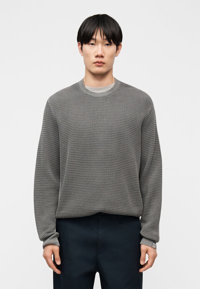 Jeune homme debout, face à l'avant, portant un pull gris texturé par-dessus une chemise gris clair et un pantalon foncé sur un fond uni.
