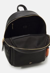Svart läderryggsäck med perforerad botten, guldzipper och innervikt. Har ett topphandtag och en tofsaccent. Modern design.