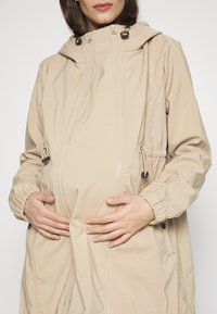 Femme enceinte portant un manteau de maternité beige, les mains posées délicatement sur son ventre.