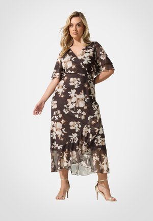 Frau trägt ein dunkel geblümtes Wickelkleid mit schwingenden Ärmeln und Rüschensaum, kombiniert mit goldfarbenen High Heels mit Knöchelriemen, steht vor weißem Hintergrund.