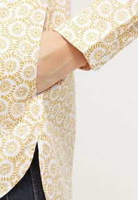 Camicia a maniche lunghe con tessuto bianco coperto da un motivo circolare giallo. Presenta polsini con bottoni e spacchi laterali all'orlo.