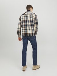 Camisa de franela a cuadros en tonos crema, azul marino y marrón; cuenta con un cuello y un diseño de botón. Combinada con jeans ajustados de color azul oscuro.