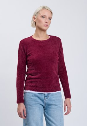 Donna con capelli biondi corti, che indossa un maglione peloso bordeaux e jeans azzurri, in piedi davanti a uno sfondo grigio chiaro uniforme.