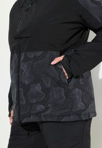 Svart jacka med en slät överdel och mönstrad nederdel som har en geometrisk design i grått. Inkluderar dragkedja fram och sidofickor.