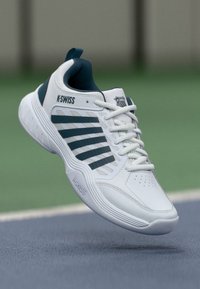 Bijela teniska obuća s zelenim detaljima, s gornjim dijelom od mrežaste tkanine, šest pruga, perforiranim dijelom za prste i teksturiranom gumom. K-Swiss brendiranje vidljivo.