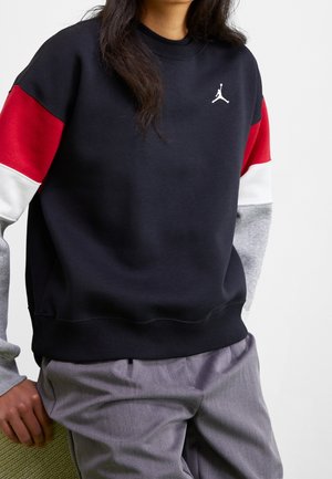 Person trägt ein farblich abgesetztes Sweatshirt mit schwarzen, roten, weißen und grauen Ärmeln und graue Hosen. Das Sweatshirt hat ein weißes Jumpman-Logo.