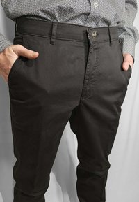 Uomo che indossa pantaloni eleganti a fantasia scura e una camicia con bottoni a fantasia, con le mani nelle tasche anteriori, su sfondo neutro.