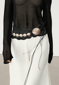 Black mesh long-sleeve top paired with white high-waisted pants, accessorized with a thin black belt featuring round beige shell accents.