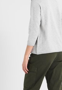 Maglione grigio chiaro a tre quarti, con una vestibilità rilassata, abbinato a pantaloni cargo verde scuro con tasche laterali. Texture liscia.