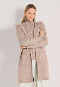 Frau trägt einen beigen Strick-Turtleneck-Pullover und dazu eine passende lange Weste mit Taschen, kombiniert mit weißen Hosen und silbernen Creolen.