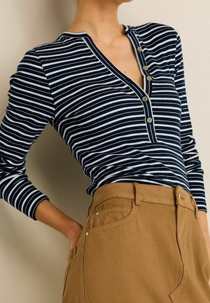 Femme portant un t-shirt henley à manches longues rayé bleu marine et blanc, rentré dans un pantalon beige taille haute, une main dans la poche.