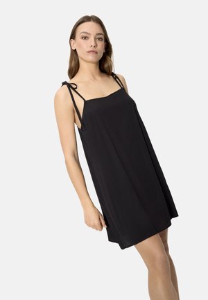 Joven mujer con un vestido negro sin mangas y tirantes anudados en los hombros, posando frente a un fondo claro y sencillo.