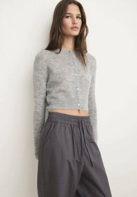 Vrouw in een lichtgrijze cropped knopen cardigan en losse donkergrijze broek met trekkoord, staande tegen een witte muur.