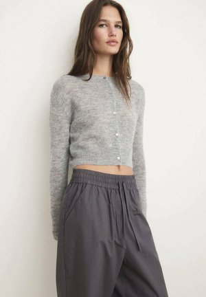 Woman wearing a light gray cropped buttoned cardigan and loose dark gray drawstring pants, standing against a white wall.