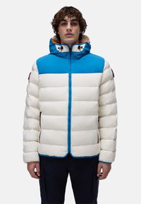 Ung man iklädd en vit och blå pufferjacka med huva och svarta dragkedjefickor, stående mot en enkel bakgrund.