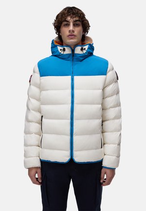 Junger Mann in einer weißen und blauen Kapuzen-Steppjacke mit schwarzen Reißverschlusstaschen, der vor einem einfarbigen Hintergrund steht.