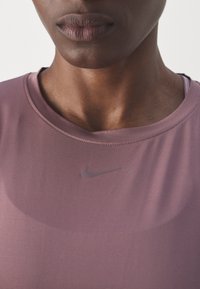 Lilla sportstrøje med rund halsudskæring, glat tekstur og et lille gråt Nike-logo foran. Nærbillede af stof og halsudskæringsdetaljer.