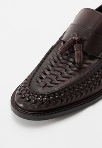 Brune læderslippers med et vævet mønster på tåen, der har et glat læderbånd og to dekorative frynser på forsiden.